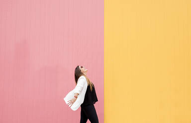 Fröhliche junge Frau mit Sonnenbrille vor rosa und gelber Wand - DAMF00702