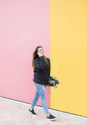 Junge Frau mit Skateboard spricht am Telefon, während sie gegen eine rosa und gelbe Wand läuft - DAMF00705
