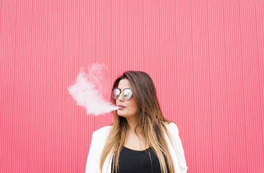Junge Frau mit Sonnenbrille beim Ausatmen von Rauch gegen eine rosa Wand - DAMF00707