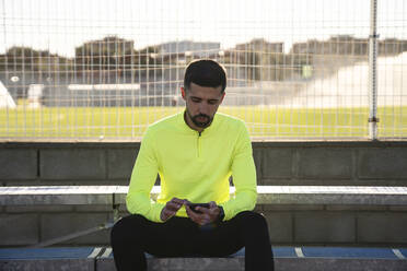 Männlicher Sportler, der sein Smartphone benutzt, während er an einem sonnigen Tag auf den Stufen eines Sportplatzes sitzt - AMPF00001