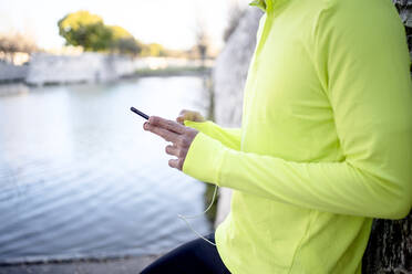 Männlicher Sportler benutzt sein Smartphone, während er in einem öffentlichen Park am See steht - AMPF00003