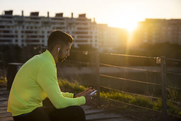 Male sportsperson listening music while sitting at park during sunset - AMPF00007