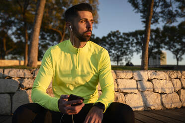 Männlicher Sportler hört Musik im Park bei Sonnenuntergang - AMPF00019