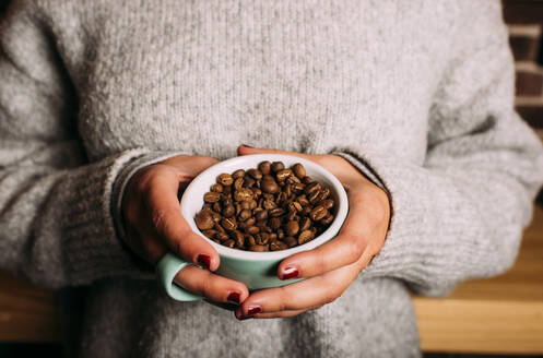 Frau mit Pullover hält Kaffeetasse mit Kaffeebohnen in der Hand - DAMF00716