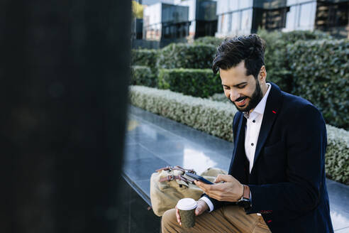 Lächelnder Geschäftsmann, der ein Mobiltelefon benutzt, während er auf einer Bank sitzt - XLGF01129