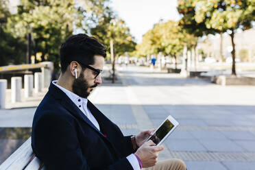 Geschäftsmann mit In-Ear-Kopfhörern, der ein digitales Tablet benutzt, während er auf einer Bank sitzt - XLGF01148