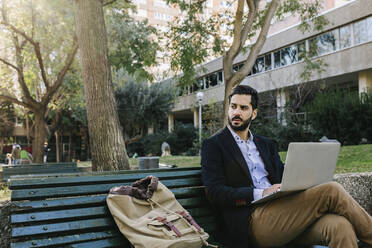 Confident businessman with laptop looking away while sitting on bench - XLGF01157