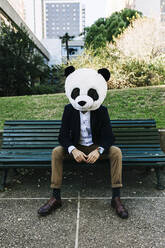 Geschäftsmann mit Panda-Maske sitzt auf einer Bank im Park - XLGF01161
