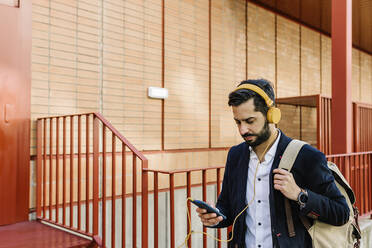 Mid adult businessman with backpack and headphones using mobile phone while standing by railing - XLGF01176