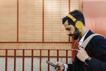 Geschäftsmann mit Kopfhörern und Rucksack, der ein Mobiltelefon benutzt, während er an einem Geländer steht - XLGF01177