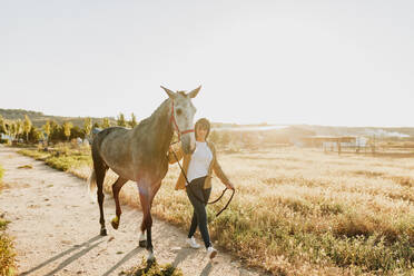 Frau, die mit ihrem Pferd auf einem Fußweg spazieren geht, an einem sonnigen Tag - MRRF00873