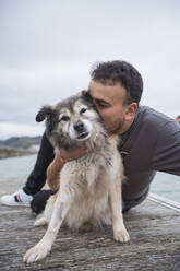 Man embracing dog while sitting on pier at beach against sky - SNF01171