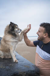 Lachender Mann gibt seinem Hund am Strand gegen den Himmel ein High Five - SNF01173