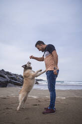 Mid adult man feeding dog while standing at beach against sky - SNF01176