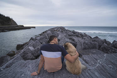 Man with arm around dog while sitting on rock against sky - SNF01178