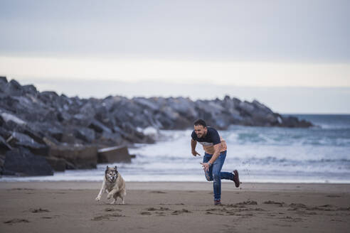 Verspielter Mann läuft mit Hund am Strand gegen den Himmel - SNF01179