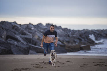 Smiling man playing with dog at beach against sky - SNF01180