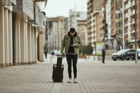 Junger Mann mit Koffer schaut auf sein Handy, während er auf einem Fußweg in der Stadt steht - ACPF01099