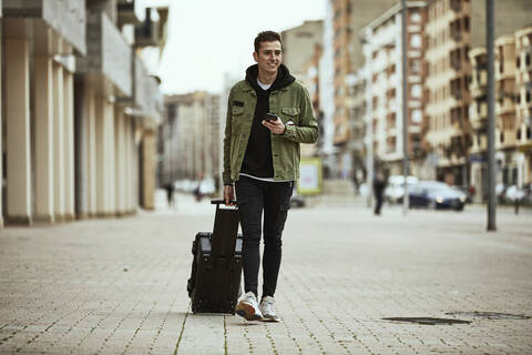 Smiling young man with suitcase holding smart phone while walking on footpath by building stock photo