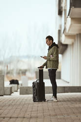 Mann mit Koffer, der sein Handy in der Hand hält, während er auf dem Fußweg vor einem Bauwerk steht - ACPF01105