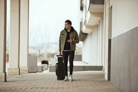 Man with suitcase holding mobile phone while looking away against sky stock photo