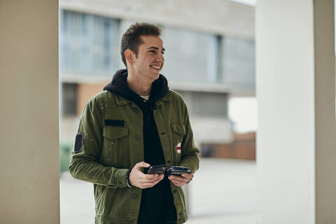 Lächelnder Mann mit Mobiltelefon, der eine Kreditkarte hält und wegschaut, lizenzfreies Stockfoto