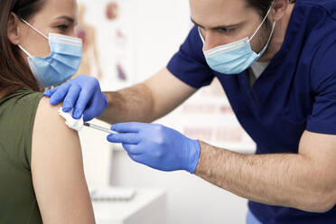 Male doctor giving vaccination to female patient at clinic during pandemic - ABIF01329