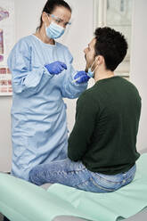 Female doctor collecting sample from mouth of male patient for COVID-19 test - ABIF01348