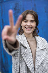 Smiling female showing peace gesture against blue wall - AXHF00161