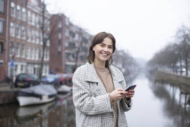 Smiling beautiful woman holding smart phone by canal against sky - AXHF00165