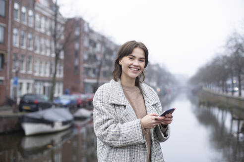 Lächelnde schöne Frau hält Smartphone am Kanal gegen den Himmel - AXHF00165