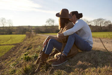 Boyfriend embracing girlfriend from behind while sitting on field during sunset - VEGF03852