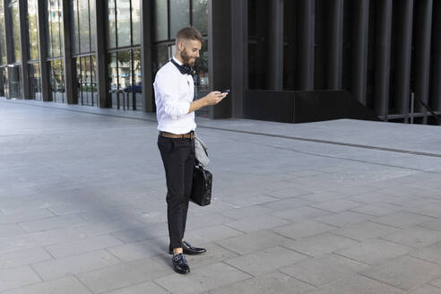 Geschäftsmann mit Aktentasche, der ein Mobiltelefon benutzt, während er auf dem Fußweg steht - BOYF01771