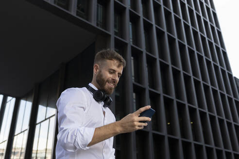 Mittlerer erwachsener Geschäftsmann, der ein Smartphone benutzt, während er an einem Gebäude steht - BOYF01772