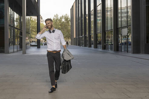 Smiling businessman with briefcase talking on mobile phone while walking on footpath - BOYF01773