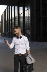 Geschäftsmann, der eine Aktentasche hält und ein Mobiltelefon benutzt, steht auf einem Fußweg - BOYF01774