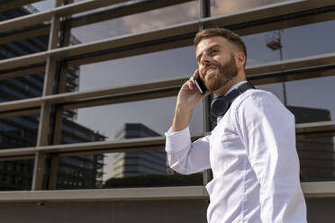 Lächelnder Geschäftsmann, der mit seinem Handy telefoniert, während er vor einem Gebäude steht - BOYF01781