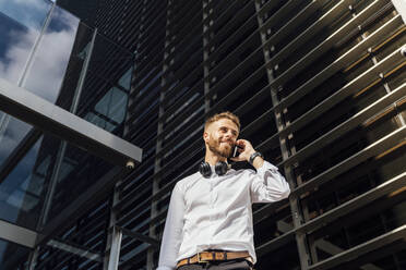 Geschäftsmann, der mit einem Mobiltelefon spricht, während er an einem Gebäude steht - BOYF01782