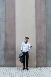 Businessman with briefcase and jacket using mobile phone while standing against wall - BOYF01787