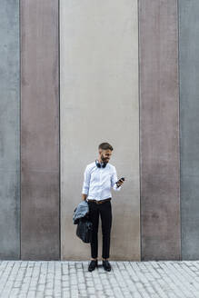 Geschäftsmann mit Aktentasche und Jacke, der ein Mobiltelefon benutzt, während er an der Wand steht - BOYF01787