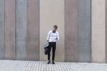 Mid adult businessman using mobile phone while standing with briefcase and jacket against wall - BOYF01790