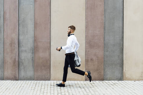 Businessman holding briefcase and jacket while running against wall - BOYF01792