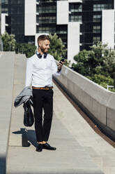 Smiling businessman using mobile phone while standing on bridge in city - BOYF01797