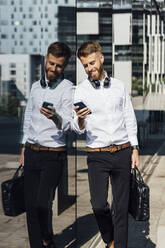 Smiling businessman with briefcase using smart phone while leaning on glass wall - BOYF01805