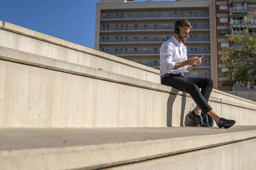 Geschäftsmann mit Kopfhörern und Smartphone, während er auf einer Treppe sitzt - BOYF01809
