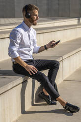 Lächelnder Geschäftsmann, der über Kopfhörer Musik hört, während er ein Mobiltelefon benutzt und auf einer Treppe sitzt - BOYF01811