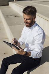 Lächelnder Geschäftsmann, der ein digitales Tablet benutzt, während er auf einer Treppe sitzt - BOYF01819