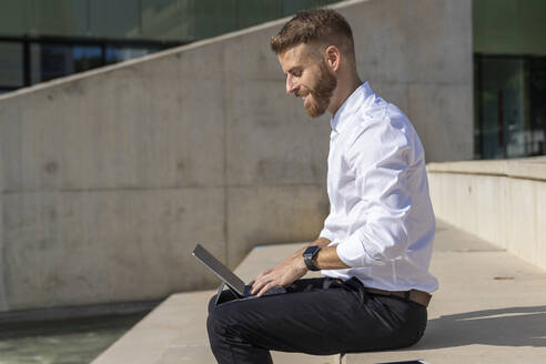 Lächelnder Unternehmer, der ein digitales Tablet benutzt, während er auf einer Treppe sitzt - BOYF01826