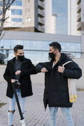 Male friends giving elbow bump wearing protective face mask while standing against buildings during COVID19 - EGAF01698