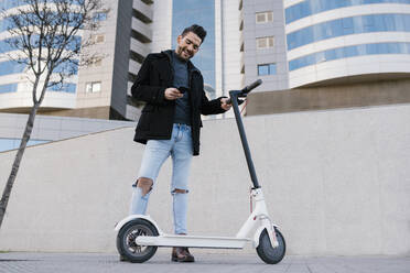 Smiling handsome man using smart phone while standing by electric push scooter against buildings - EGAF01699